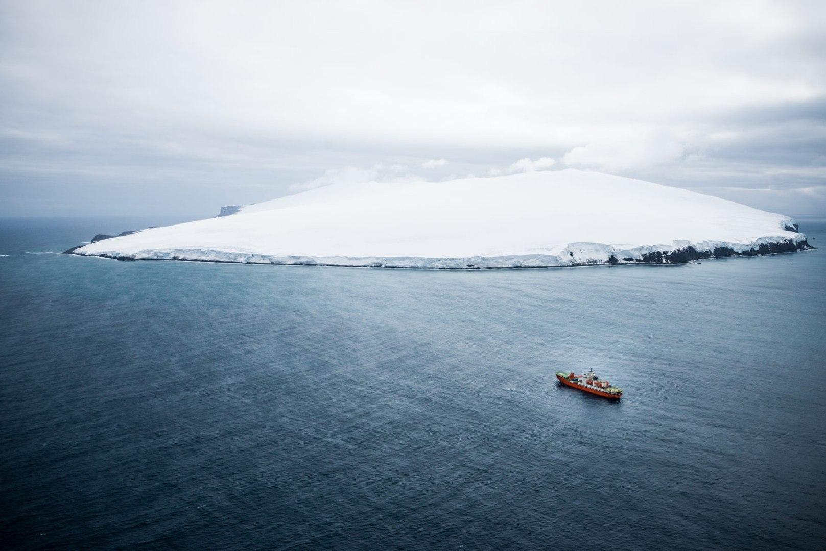 Vue aérienne de l’île Bouvet entièrement enneigée, avec un navire de recherche au large